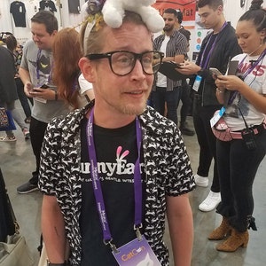 May include: A person wearing a white cat plushie on their head, a black and white patterned shirt, and a black t-shirt with the text "Bunny Ears". They are wearing glasses and a CatCon Talent badge.
