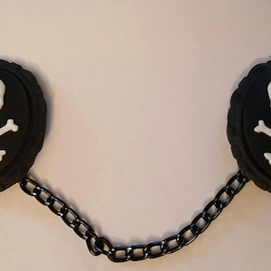 May include: A pair of black skull and crossbones toggle clasps connected by a black chain. Each clasp features a white skull and crossbones design.