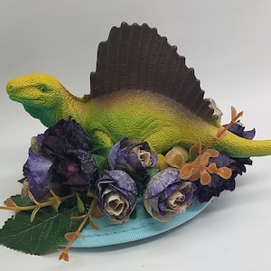May include: A green and yellow dinosaur figurine with a brown sail fin, surrounded by purple and brown flowers, on a blue base.