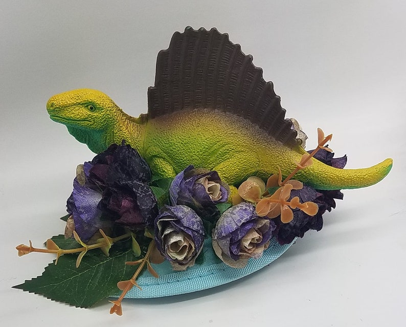 May include: A green and yellow dinosaur figurine with a brown sail fin, surrounded by purple and brown flowers, on a blue base.