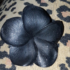 May include: A black glitter plumeria flower. The flower has five petals and is made of a soft, fabric-like material.