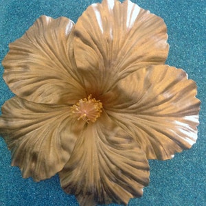 May include: A large, golden hibiscus flower with a textured surface. The flower has five petals and a center with a light pink pistil.