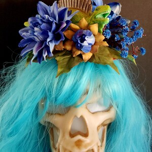 May include: A blue and yellow floral crown with a green dinosaur figurine on top. The crown is worn on a blue wig over a white skull mask.
