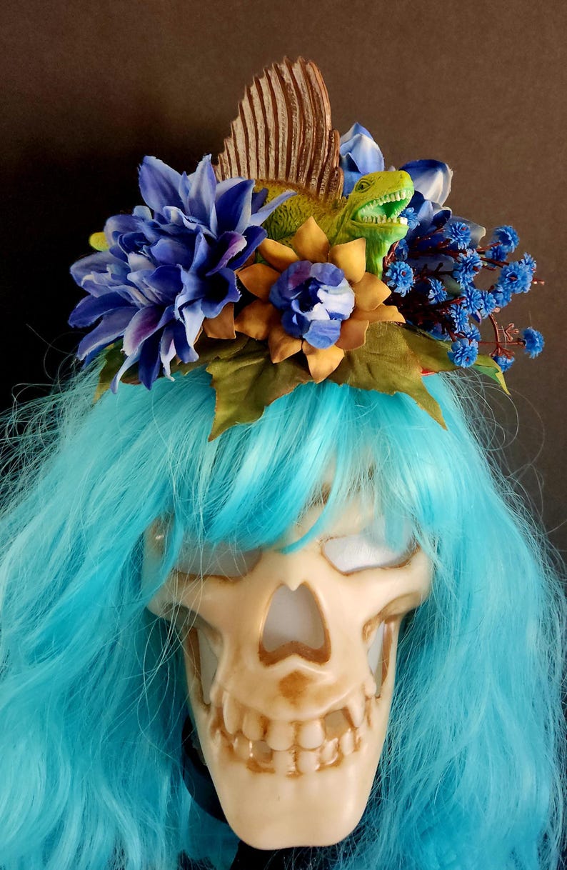 May include: A blue and yellow floral crown with a green dinosaur figurine on top. The crown is worn on a blue wig over a white skull mask.
