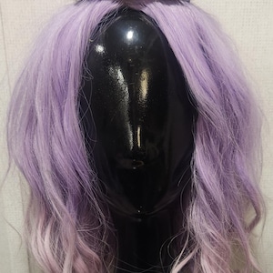 Puede incluir: Un tocado negro con dos cuernos curvados se asienta sobre una cabeza de maniquí con cabello ondulado color lavanda. Los cuernos tienen una apariencia texturizada. La peluca tiene una onda natural.