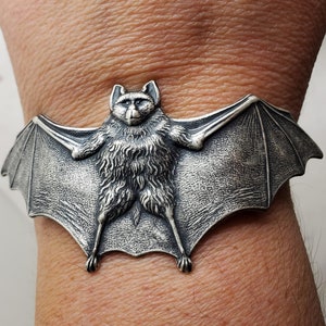 May include: A silver bat bracelet with a detailed, textured design. The bat is depicted in a flying position with its wings spread wide.