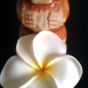 May include: A white and yellow plumeria flower rests in front of a brown tiki statue. The flower has five rounded petals. The tiki statue has a carved face and is brown with orange accents.