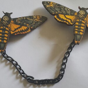 May include: A pair of black chain connected  moth brooches with a skull design. The moths are painted in shades of green, yellow, and black.
