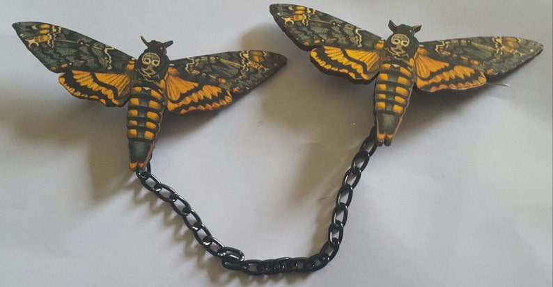 May include: A pair of black chain connected  moth brooches with a skull design. The moths are painted in shades of green, yellow, and black.