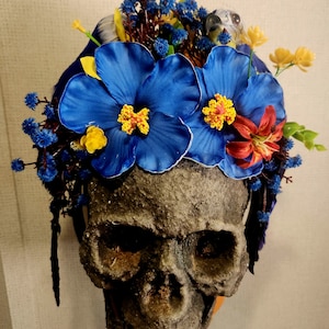 May include: A skull with a floral crown made of blue, yellow, and red flowers. The crown is made of silk flowers and has a blue ribbon tied around it.