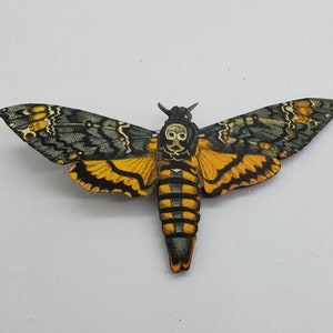 May include: A black and yellow death's-head hawkmoth brooch with a skull design on its thorax. The moth is depicted with intricate details and realistic colors.