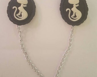 May include: A pair of black and white cat cameo brooches connected by a silver chain. Each brooch features a white cat silhouette on a black background.