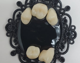 May include: A black, ornate, oval-shaped frame with a glossy black center. The frame holds several realistic-looking, off-white teeth. The frame has a detailed, vintage design with decorative elements.