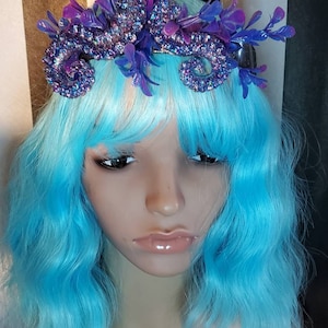May include: A purple and blue glitter octopus hair crown with purple and blue plastic flowers. The crown is on a blue wig.