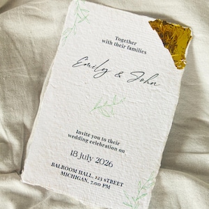 Puede incluir: Una invitación de boda blanca y texturizada con los nombres "Emily & John" en escritura elegante. La invitación incluye detalles para la celebración de la boda el 18 de julio de 2026, en Michigan. Ilustraciones de hojas verdes y detalles dorados.