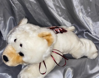 May include: A white plush toy in the shape of a dog lying on its belly with its head turned to the side. The dog has a red ribbon around its neck and a red and white striped pattern on its back.
