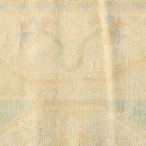 Op de afbeelding: Een geweven textielkleed met een lichtbeige basiskleur en een herhalend patroon van zachtblauwe en gedempte oranje bloemmotieven. De stof vertoont een zichtbare rasterachtige weefstructuur over het gehele oppervlak met geometrische randelementen.