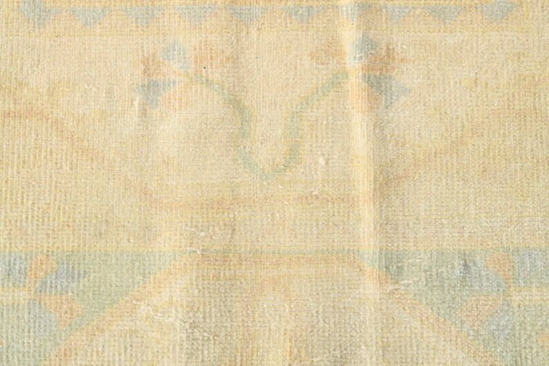 Op de afbeelding: Een geweven textielkleed met een lichtbeige basiskleur en een herhalend patroon van zachtblauwe en gedempte oranje bloemmotieven. De stof vertoont een zichtbare rasterachtige weefstructuur over het gehele oppervlak met geometrische randelementen.