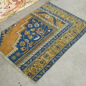 May include: A rectangular, vintage rug with a detailed pattern. The rug features a central blue field with floral motifs, bordered by a yellow and brown design. The rug's edges are finished with a fringe.