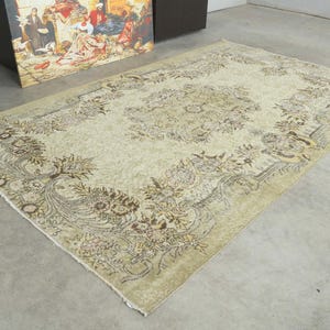 May include: A rectangular area rug featuring a light beige background with a central floral medallion and intricate botanical borders in shades of brown, tan, and muted gold. The rug has a low pile texture and short fringe along the edges.