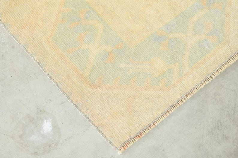 Op de afbeelding: Een geweven kleed met een lichtbeige veld en een zachte saliegroene rand. Het kleed toont een herhalend geometrisch patroon in gedempte oranje en cr&egrave;metinten, afgewerkt met een korte, gefranjerde rand langs de zijkant van het rechthoekige textiel.