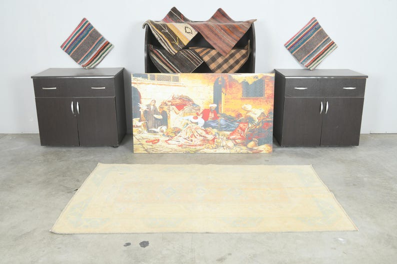 Op de afbeelding: Een rechthoekig lichtbeige vloerkleed met een subtiel geometrisch randpatroon, geplaatst voor twee donkerbruine kasten, een zwarte kast met gevouwen textiel met patronen en een groot rechthoekig schilderij met een markttafereel.