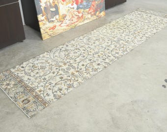May include: A long, rectangular runner rug featuring a cream-colored base with an intricate, repeating floral and vine pattern in shades of muted blue, brown, and gray. The rug has a distinct border at one end with a dense, detailed botanical design.