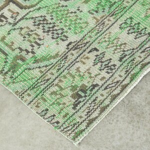 May include: A woven rug featuring a detailed pattern in shades of green, cream, and dark brown. The design includes intricate geometric motifs and a decorative border with repeating shapes, showcasing a textured surface and a finished edge.