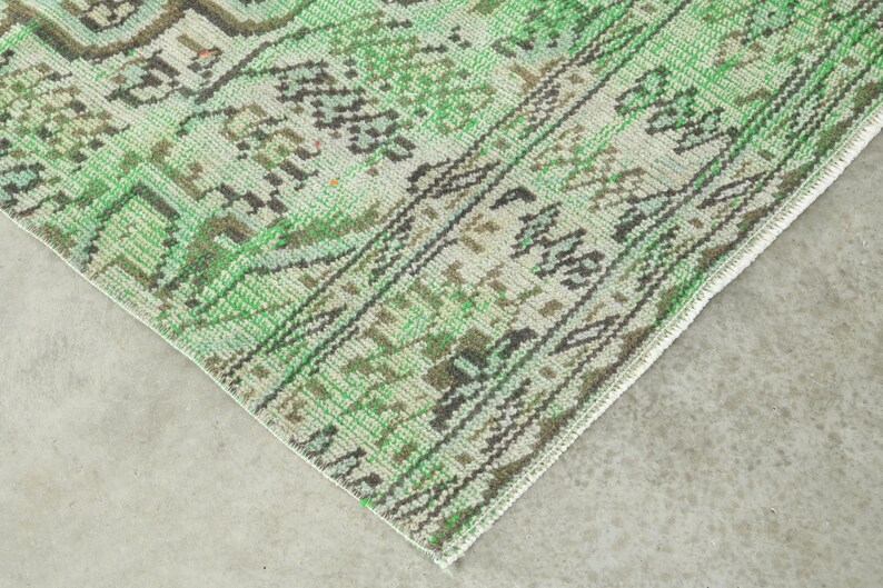 May include: A woven rug featuring a detailed pattern in shades of green, cream, and dark brown. The design includes intricate geometric motifs and a decorative border with repeating shapes, showcasing a textured surface and a finished edge.