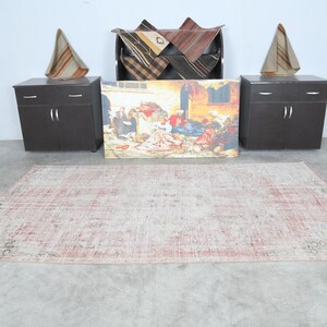 May include: A light pink and cream coloured vintage rug with a distressed look. The rug is laid out on a concrete floor in a room with two dark brown cabinets and a framed painting of people in a market scene.