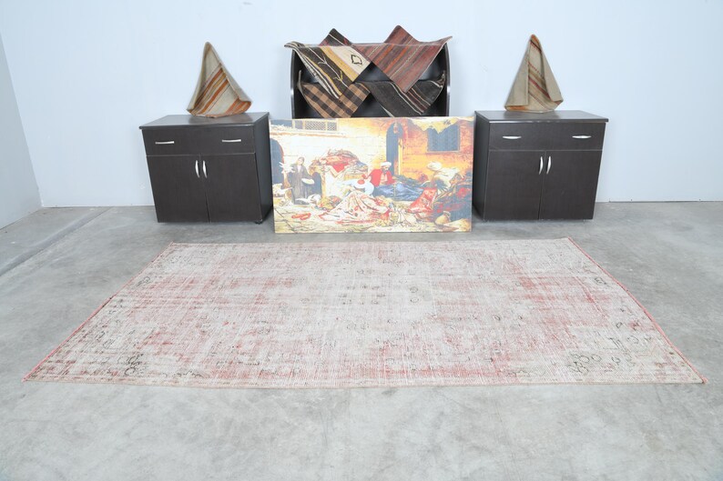 May include: A light pink and cream coloured vintage rug with a distressed look. The rug is laid out on a concrete floor in a room with two dark brown cabinets and a framed painting of people in a market scene.