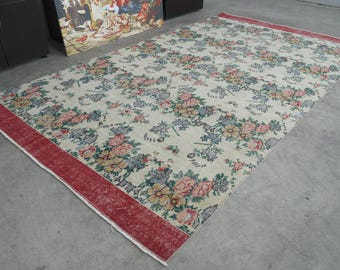 May include: Rectangular floral rug with a cream background and a red border. The rug features a repeating pattern of pink, blue, and yellow flowers and green leaves. The rug is placed on a gray concrete floor.