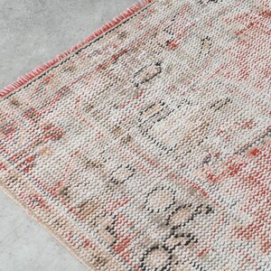 May include: A vintage rug with a cream background and a red and black geometric pattern. The rug has a fringed border.