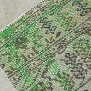 May include: A woven rug featuring a vibrant green and muted sage color palette with dark brown geometric and floral patterns. The textile displays a detailed, repeating motif across its surface, finished with a clean, straight white fringe edge.
