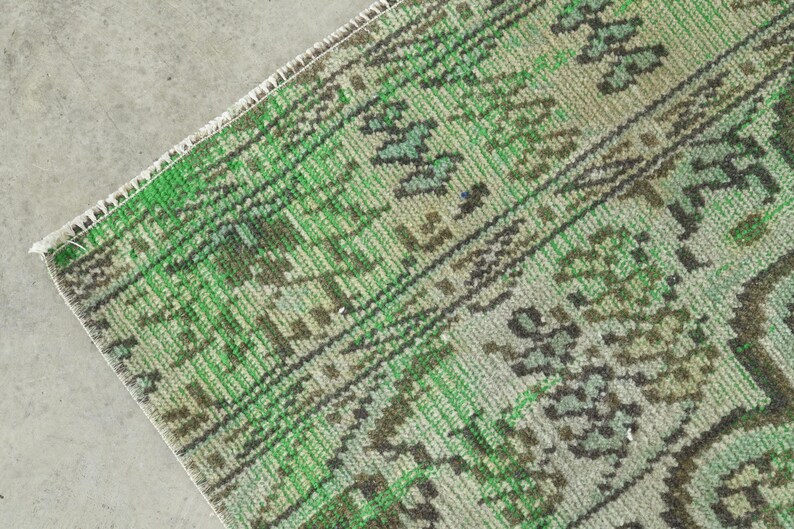 May include: A woven rug featuring a vibrant green and muted sage color palette with dark brown geometric and floral patterns. The textile displays a detailed, repeating motif across its surface, finished with a clean, straight white fringe edge.