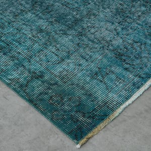May include: Close-up of a teal blue rug with a faded pattern. The rug has a white fringe border.