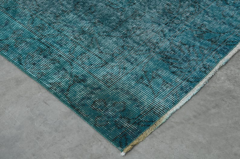 May include: Close-up of a teal blue rug with a faded pattern. The rug has a white fringe border.