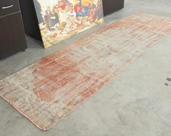 Può includere: Un lungo tappeto passatoia rettangolare caratterizzato da un motivo sbiadito e usurato nei toni dell'arancione terracotta, beige e crema. Il tappeto presenta una texture invecchiata con segni visibili di usura e una struttura a tessitura piatta.