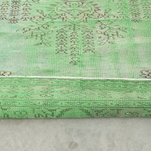 May include: A rectangular area rug featuring a vibrant green base color with intricate, faded brown geometric and floral patterns. The rug is partially folded over, revealing a textured weave and a distinct border design along the edges.