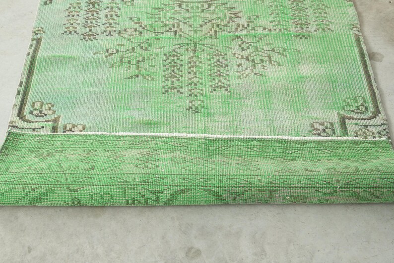 May include: A rectangular area rug featuring a vibrant green base color with intricate, faded brown geometric and floral patterns. The rug is partially folded over, revealing a textured weave and a distinct border design along the edges.