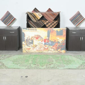 May include: A rectangular green rug with a central medallion and intricate border patterns, two dark brown cabinets with silver handles, two striped square pillows, a dark storage unit with folded textiles, and a rectangular painting of a scene.