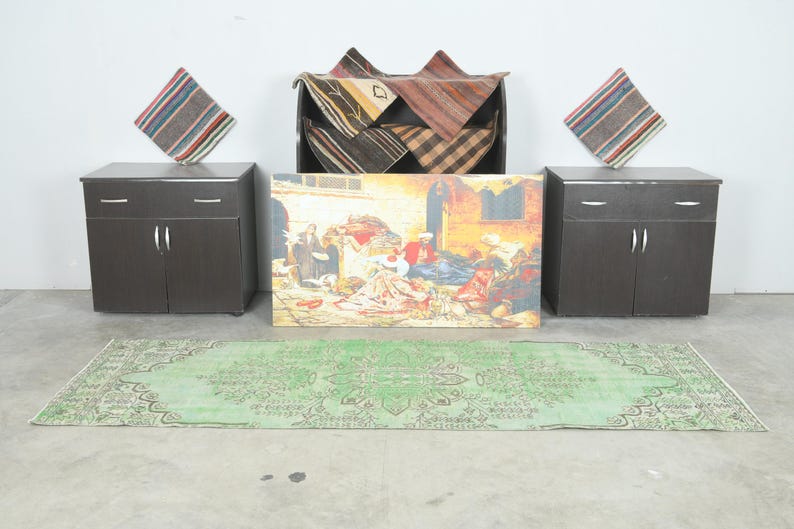 May include: A rectangular green rug with a central medallion and intricate border patterns, two dark brown cabinets with silver handles, two striped square pillows, a dark storage unit with folded textiles, and a rectangular painting of a scene.
