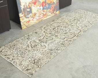 May include: A long, rectangular runner rug featuring a faded, distressed pattern in shades of beige, cream, and dark brown. The design includes intricate central medallions and a repeating geometric border motif across the entire surface.