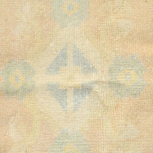 Op de afbeelding: Een geweven textielkleed met een herhalend patroon van lichtblauwe en gele bloemmotieven op een getextureerde beige achtergrond. Het ontwerp bevat geometrische ruitvormen afgewisseld met gestileerde bloemelementen over het oppervlak.