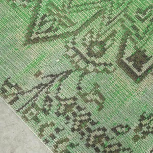 May include: A woven rug featuring a vibrant green base color with intricate dark brown geometric and floral patterns. The textile displays a textured surface with a visible weave, showcasing a repeating symmetrical design across the fabric.