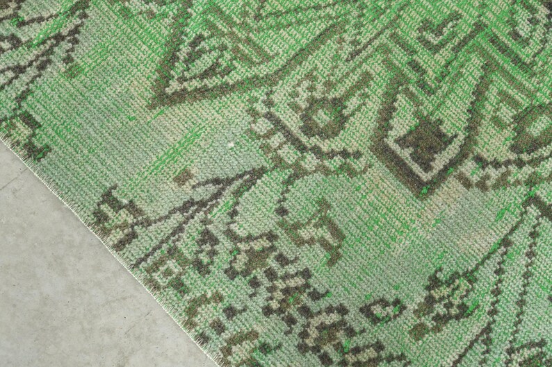 May include: A woven rug featuring a vibrant green base color with intricate dark brown geometric and floral patterns. The textile displays a textured surface with a visible weave, showcasing a repeating symmetrical design across the fabric.