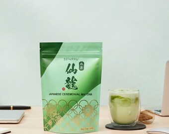 Puede incluir: Una bolsa verde de Senryu Japanese Ceremonial Matcha con detalles dorados y un vaso de matcha latte sobre un escritorio de madera. El matcha está en un vaso transparente con un posavasos negro.