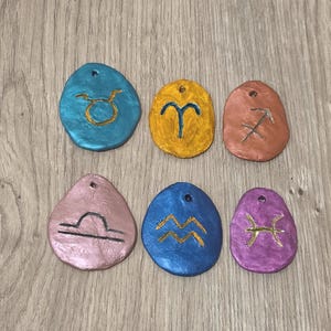 Könnte beinhalten: Eine Sammlung von sechs farbenfrohen, oval geformten Anhängern, die jeweils ein anderes Tierkreiszeichen zeigen. Die Anhänger sind in Blau-, Gold-, Orange-, Rosa- und Lilatönen gehalten und haben goldene oder schwarze Symbole. Jeder hat ein kleines Loch zum Aufhängen.