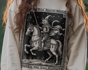 Op de afbeelding: Beige sweatshirt met een zwart-wit illustratie van een kattenridder te paard, met de tekst "Felis Ingratus" en "The Nine Lives Oath". De sweatshirt heeft lange mouwen en een ronde hals.
