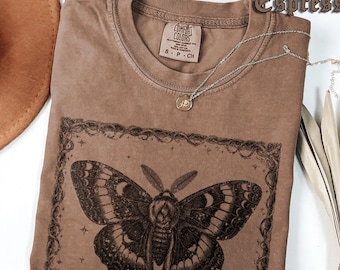 May include: Espresso-colored t-shirt with a large moth graphic in a decorative frame. The shirt has a crew neck and short sleeves. A silver necklace with a small pendant rests on the shirt. The shirt is made of a soft material.